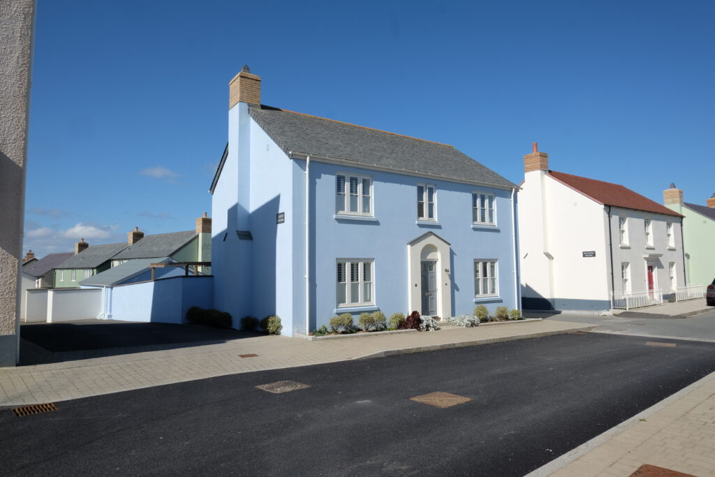 A two-story light blue house with white trim and a gray roof stands on a sunny residential street, surrounded by similar houses. The sky is clear and blue, and the road in front is freshly paved.