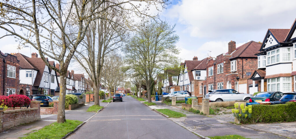 A quiet suburban street lined with trees and parked cars, flanked by semi-detached brick houses with front gardens under a partly cloudy sky.