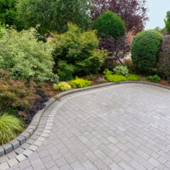 A neatly paved driveway curves alongside a well-maintained garden bed filled with various green shrubs, bushes, and small trees. The landscaping appears lush and tidy.