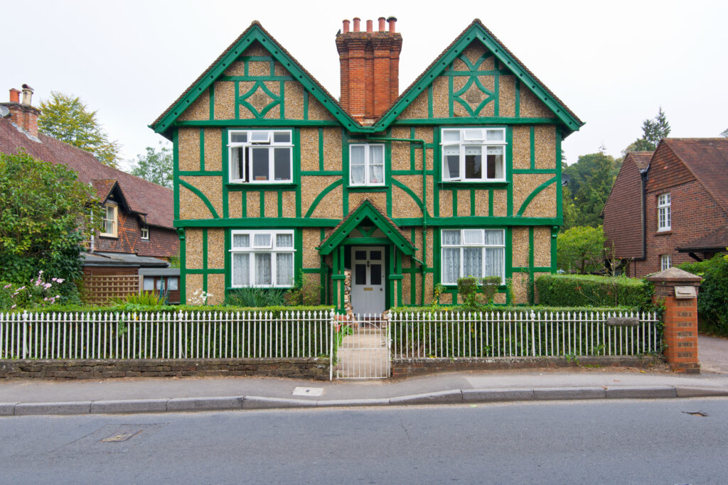 A two-story house with decorative green timber framing and beige walls, a central porch, white-framed windows, and three red brick chimneys. The house is surrounded by a white picket fence and greenery.