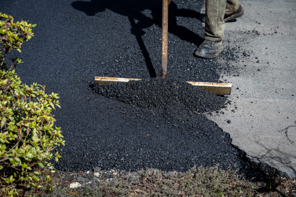 A person uses a rake to spread fresh asphalt on a driveway, with green bushes nearby and only the lower legs visible.