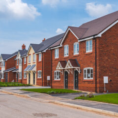 A row of modern, red-brick houses with small front lawns and driveways on a quiet, newly built suburban street under a blue sky with scattered clouds.