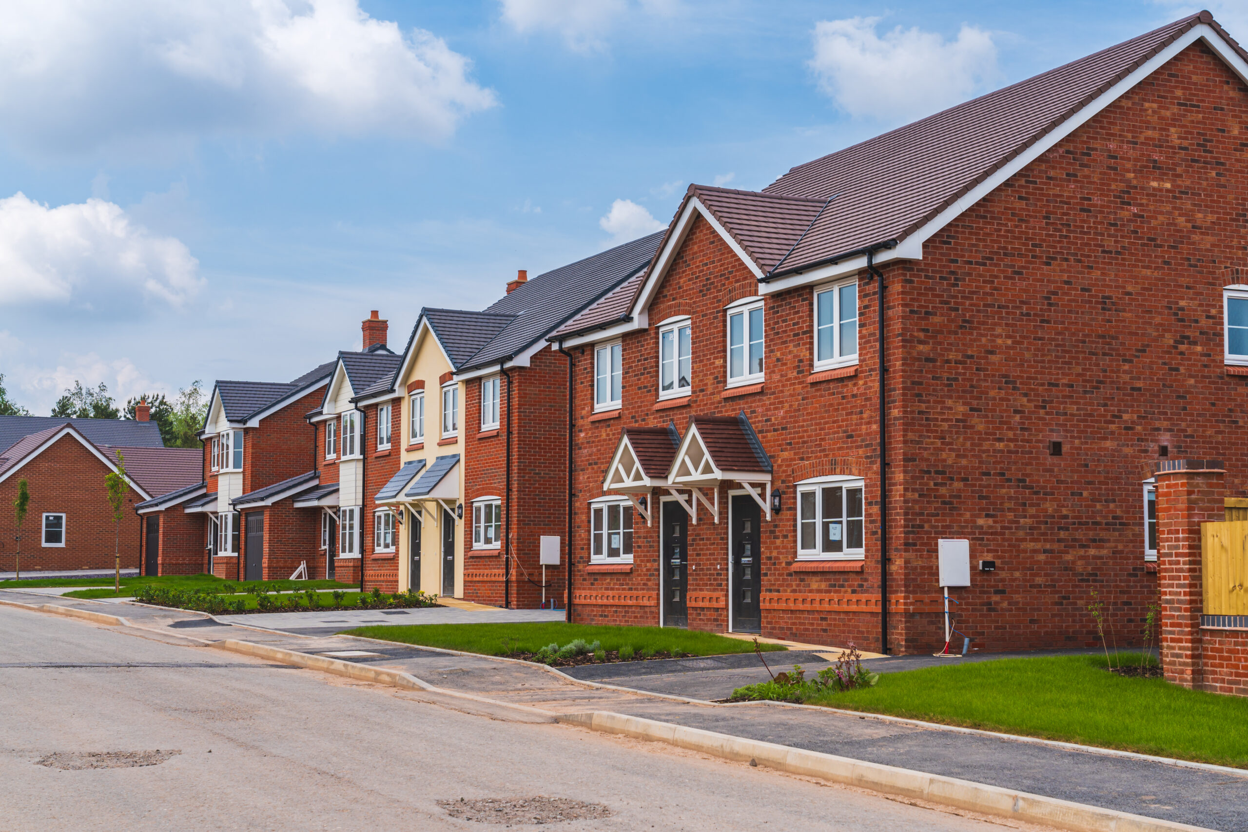 A row of modern, red-brick houses with small front lawns and driveways on a quiet, newly built suburban street under a blue sky with scattered clouds.