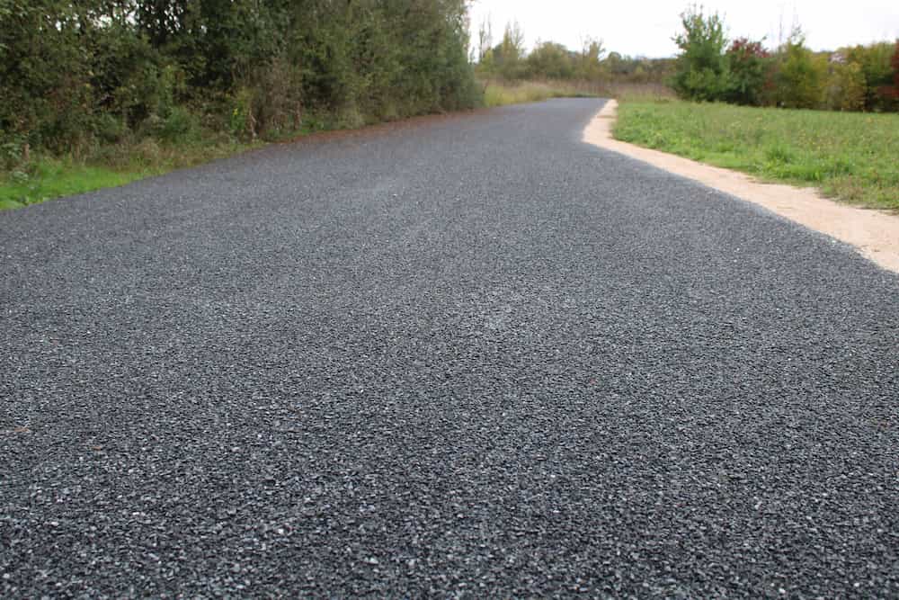 A newly paved asphalt road curves through a green, grassy area, bordered by trees and bushes on one side, under a cloudy sky.