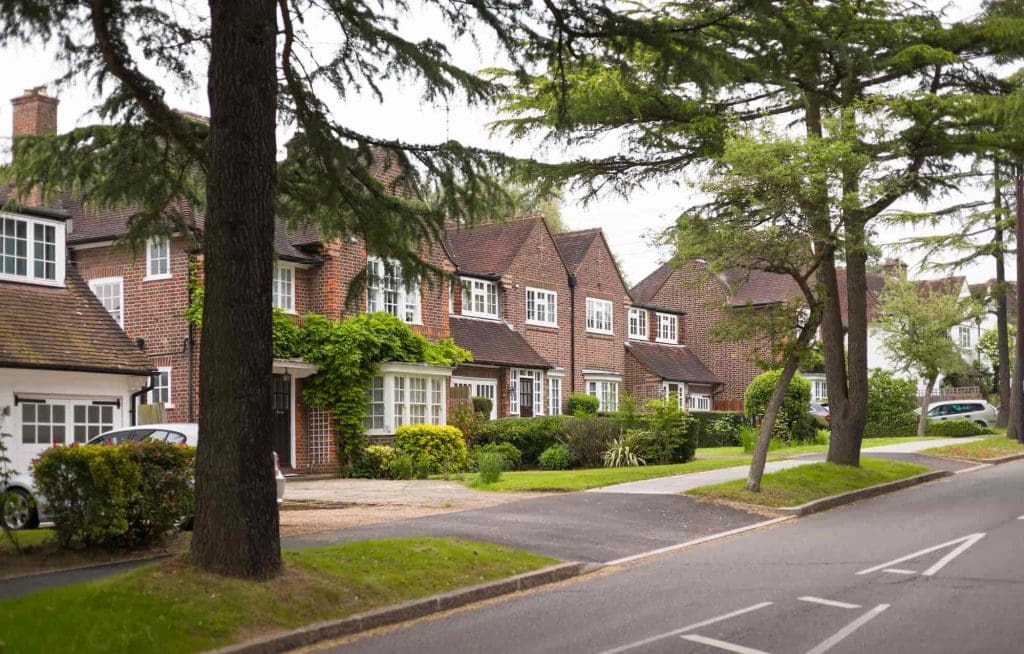 A row of brick houses with tiled roofs, large windows, and well-kept front gardens lines a quiet suburban street bordered by trees and a sidewalk.