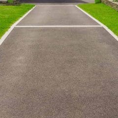 A smooth, freshly-paved driveway bordered by white bricks, with green grass and bushes on one side and a stone wall with plants on the other, leading toward a home.