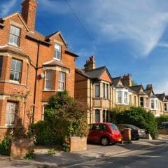A row of traditional brick and stucco houses with bay windows lines a quiet residential street. Several cars are parked along the curb, and greenery and shrubs decorate the front gardens under a blue sky.