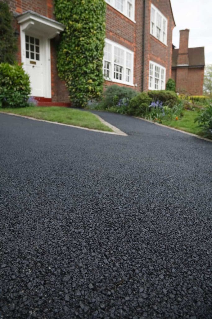 A smooth, freshly paved black asphalt driveway leads up to a red-brick house with white windows and a white front door, surrounded by green plants and shrubs.