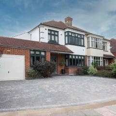 A two-story house with a white and red brick exterior, black window frames, a bay window, attached garage, and a spacious paved driveway surrounded by shrubs and greenery under a partly cloudy sky.