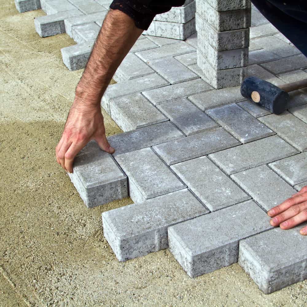 A person is laying rectangular gray paving stones in a herringbone pattern on a sandy surface, using their hands to position the bricks and a rubber mallet nearby.