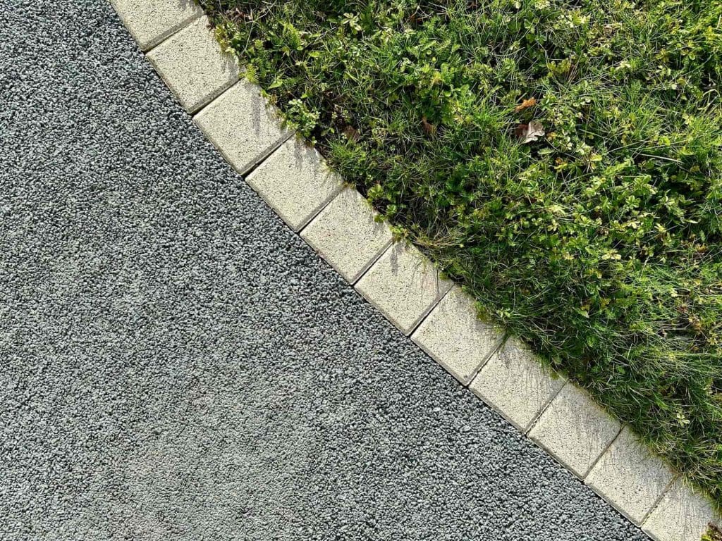 A curved stone border separates a gravel path on the left from green grass with small plants on the right. The photo is taken from above, showing the distinct textures and colors of each section.