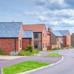 A row of modern brick houses with slate roofs lines a curved residential street, bordered by green lawns and gardens under a partly cloudy sky.