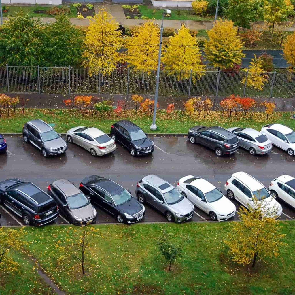 Aerial view of a parking lot with two rows of cars, surrounded by trees with yellow and green autumn foliage. The cars are parked on wet pavement, suggesting recent rain. A sidewalk and road are visible in the background.